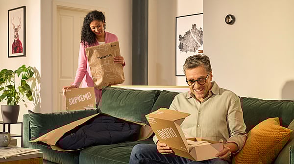 Bir adam, önünde bir Protective Mailer bulunan bir Mondi DelightBox'ı açarken kanepeye oturuyor, arkasında ise bir kadın MailerBag ve SupremeBox tutarak ayakta duruyor.