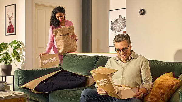 Bir adam, önünde bir Protective Mailer bulunan bir Mondi DelightBox'ı açarken kanepeye oturuyor, arkasında ise bir kadın MailerBag ve SupremeBox tutarak ayakta duruyor.