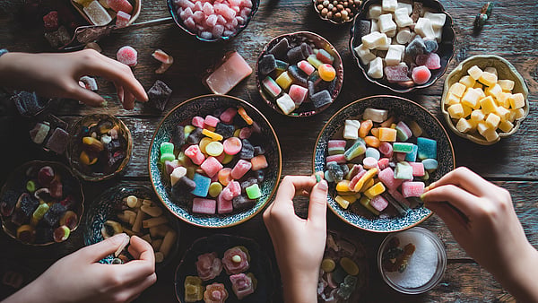 Ahşap bir masanın üzerinde, renkli şekerlemeler, marshmallowlar ve tatlılarla dolu kaselere uzanan birkaç elin yukarıdan görünümü. Masanın etrafına, çeşitli şekil, renk ve dokulara sahip şekerlemelerin bulunduğu çok sayıda tabak dizilmiş; bu da ortak bir atıştırma sahnesi oluşturuyor.